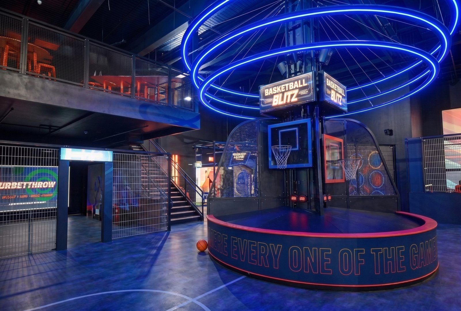 Design and engineering — a neon-lit basketball attraction on a raised podium beneath suspended light rings, with a steel mezzanine, mesh balustrades and a games hall beyond
